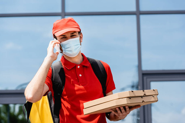Kurier in medizinischer Maske spricht mit Smartphone und hält Pizzakartons auf der Straße  - Foto, Bild