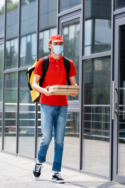 Repartidor en máscara médica con mochila térmica sosteniendo cajas de pizza mientras camina por la calle urbana
  - Foto, Imagen