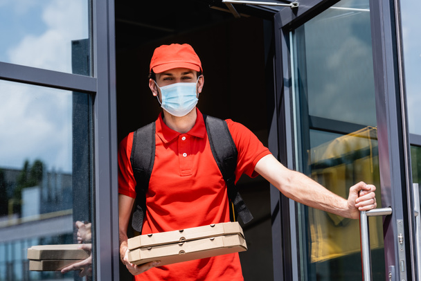 Leverancier in medisch masker en uniform met pizzadozen in de buurt van open deur van het gebouw  - Foto, afbeelding