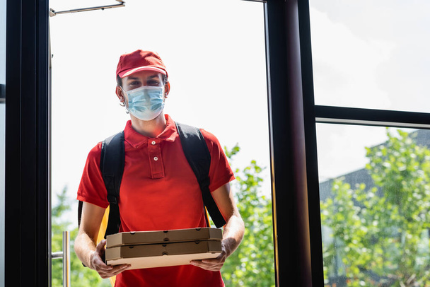 Courrier dans un masque médical tenant des boîtes à pizza près de la porte ouverte du bâtiment  - Photo, image