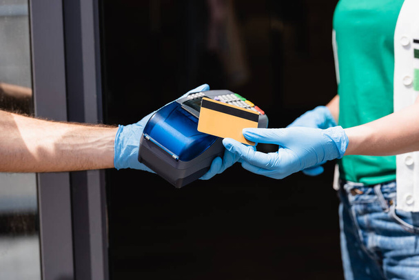 Gewassen beeld van vrouw in latex handschoenen met creditcard in de buurt van koerier met betaalterminal op stedelijke straat  - Foto, afbeelding