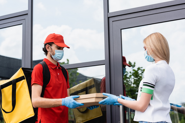 Courier in medical mask and latex gloves giving pizza boxes to woman near building on urban street  - 写真・画像