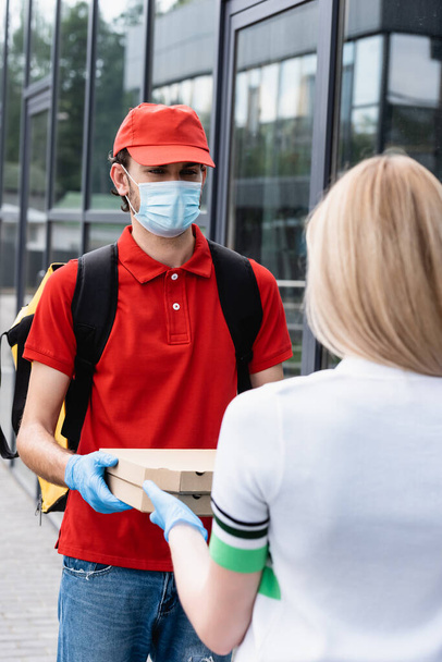 Selective focus of courier in uniform and medical mask giving takeaway pizza to woman on urban street   - Fotoğraf, Görsel