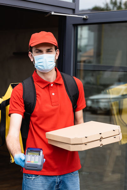 Courier in medical mask and latex glove holding payment terminal and pizza boxes on urban street  - Photo, Image