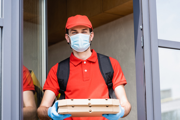 Delivery man in medical mask and latex gloves holding pizza boxes near building on urban street  - Fotó, kép