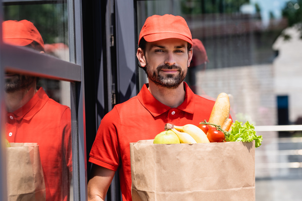 Handsome delivery man holding shopping bag with grocery on urban street  - 写真・画像