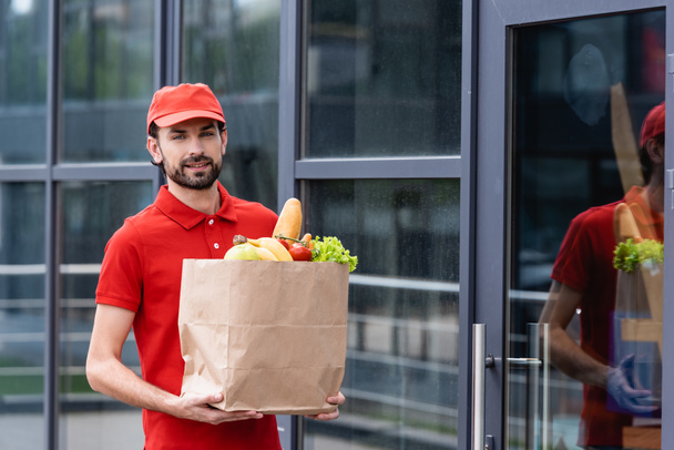 Guapo repartidor hombre sosteniendo bolsa de compras con frutas y verduras frescas en la calle urbana
  - Foto, imagen