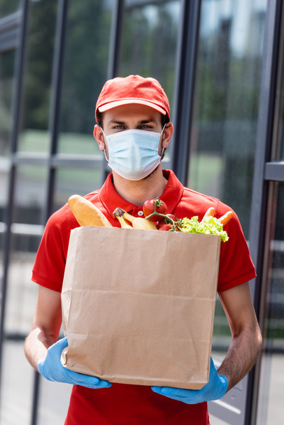 Доставщик в медицинской маске и латексных перчатках держит сумку с продуктами на городской улице  - Фото, изображение