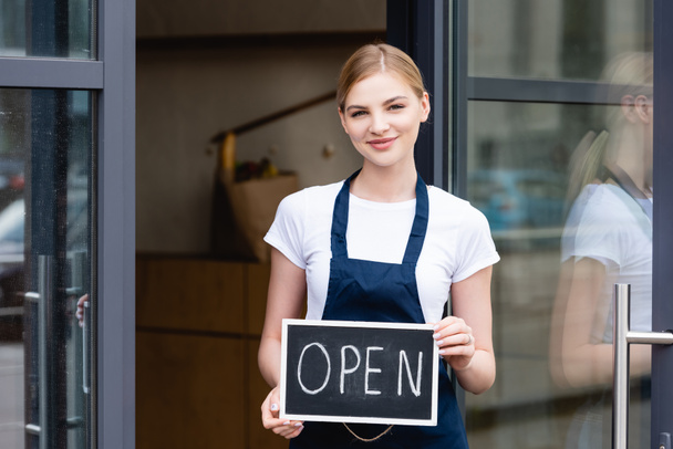 Smiling waitress holding signboard with open lettering near door of cafe on urban street  - Photo, Image
