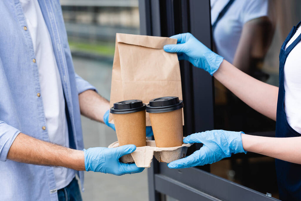 Cropped view of man receiving paper cups and package from waitress in latex gloves near cafe  - 写真・画像