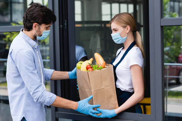 Vista lateral del hombre tomando bolsa de compras con frutas y verduras de la vendedora cerca de la ventana
 - Foto, imagen