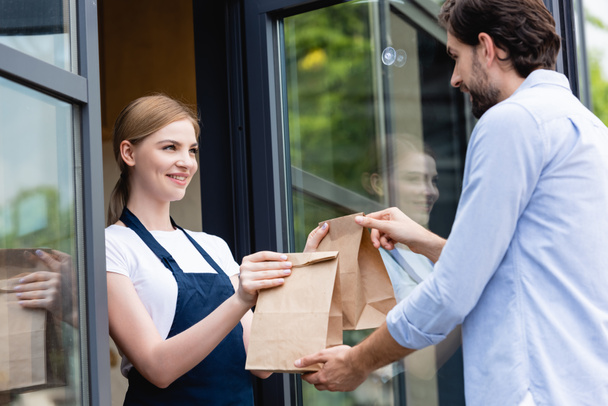 Attractive saleswoman giving paper bags to man near door of cafe - Photo, Image