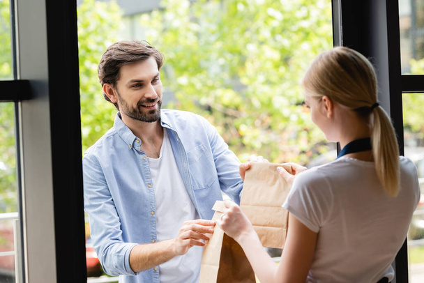 Selective focus of handsome man receiving packages from waitress at entry of cafe - Photo, image