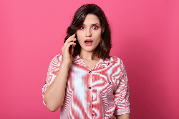 Shocked lady with widely opened mouth talking to phone, hears shocking news, being very surprised, wears shirt, has dark hair, standing against pink wall. - Photo, image