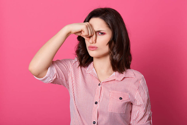 Caucasian women dresses striped shirt feeling bed smell, keeping fingers on her nose, suffering from stinking air, posing against pink wall, standing with unpleasant facial expression. - Foto, afbeelding