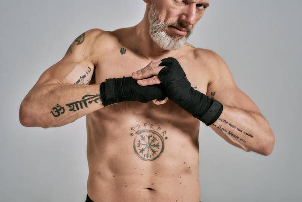Homem atlético de meia-idade meio nu, kickboxer aquecendo as mãos envoltas antes de treinar de pé em estúdio sobre fundo cinza
 - Foto, Imagem