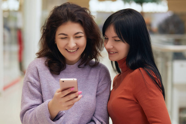 Friends having fun with smart phone, looking at mobile phones screen with happy expression and laughing, females wearing sweaters, posing together while reading social network. - Foto, Bild