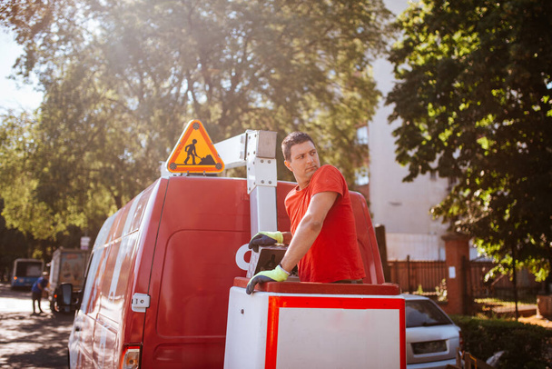  A caucasian worker in a red T-shirt stands in a basket in a crane . Setting up traffic lights in progress - Fotoğraf, Görsel