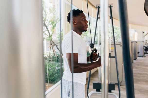 Muscular African American sportsman doing cable fly with exercise machine standing against window during training in gym - Φωτογραφία, εικόνα