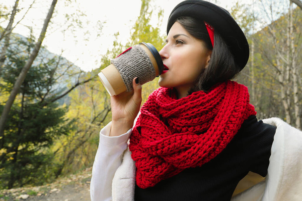 Hermosa mujer joven en un sombrero negro bebe café de la taza acogedora en la naturaleza. Picnic al aire libre buen humor
. - Foto, imagen