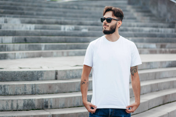 A young stylish man with a beard in a white T-shirt and glasses. Street photo - Foto, Bild