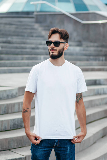 A young stylish man with a beard in a white T-shirt and glasses. Street photo - Foto, afbeelding