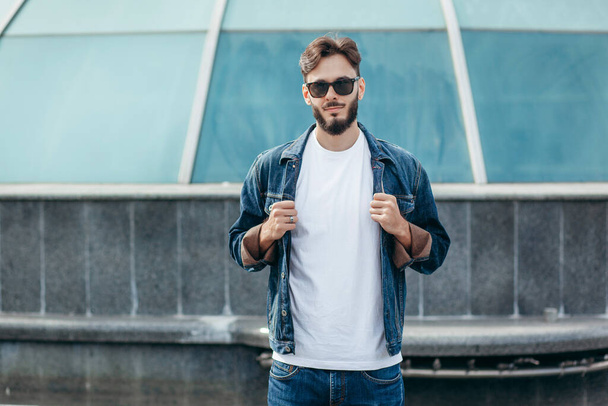 A young stylish man with a beard in a white T-shirt and glasses. Street photo - Photo, Image