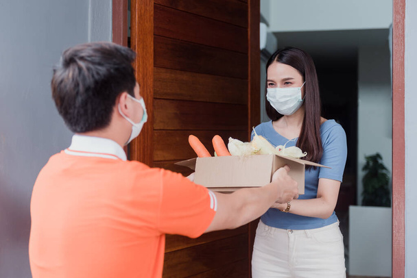 アジアの男性はオレンジのシャツを着て配達製品を運ぶ。野菜や果物を入れた紙箱を自宅のアジア人女性に送る. - 写真・画像