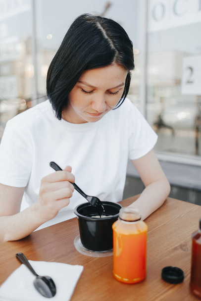 A young woman with black hair in a white T-shirt sits on an open veranda in a cafe and eats food from disposable plastic dishes.  - Foto, afbeelding