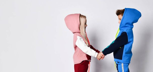 Little kids, brother and sister, in hoods, colorful tracksuits and sneakers. They holding hands, posing sideways isolated on white - Photo, image
