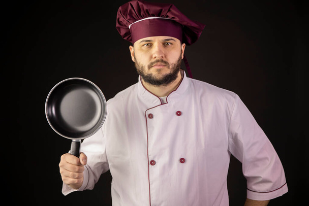Майстер шеф-кухар в уніформі руків'я сковорідка дивиться на камеру ізольовані на чорному
 - Фото, зображення