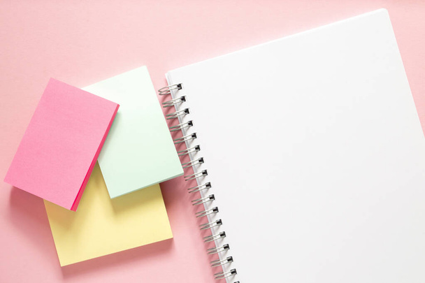 Λευκό σημειωματάριο με σπείρα και τρία πολύχρωμα αυτοκόλλητα σε ένα λεπτό ροζ φόντο. Διάταξη και επίπεδη θέσει με θέση για κείμενο. Αντίληψη πίσω στο σχολείο. - Φωτογραφία, εικόνα