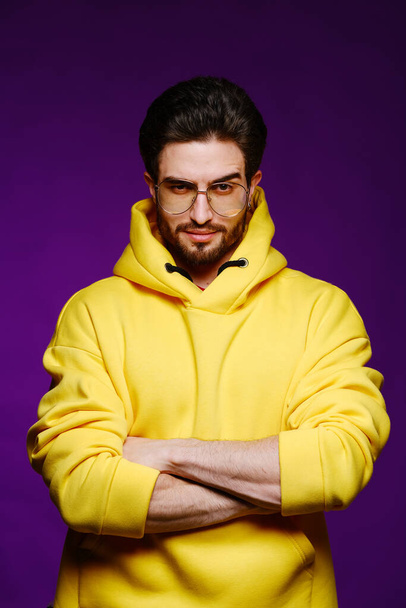 A young man of 25-30 years in glasses and a yellow sweatshirt emotionally poses on a purple background.  - Fotó, kép