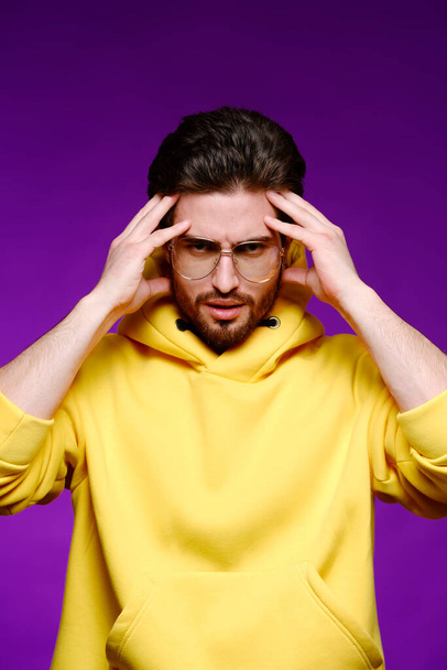 A young man of 25-30 years in glasses and a yellow sweatshirt emotionally poses holding his head on a purple background. - Photo, Image