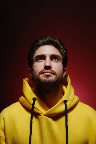 Portrait of a young guy 25-30 years old looking up in a yellow sweatshirt on red wall background.  - Fotó, kép