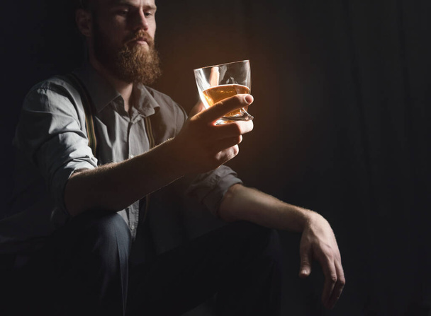 Гарний молодий чоловік з бородою сидить у темній кімнаті зі склянкою алкоголю в руках. Поняття відпочинку і релаксації, бузку і самотності
 - Фото, зображення