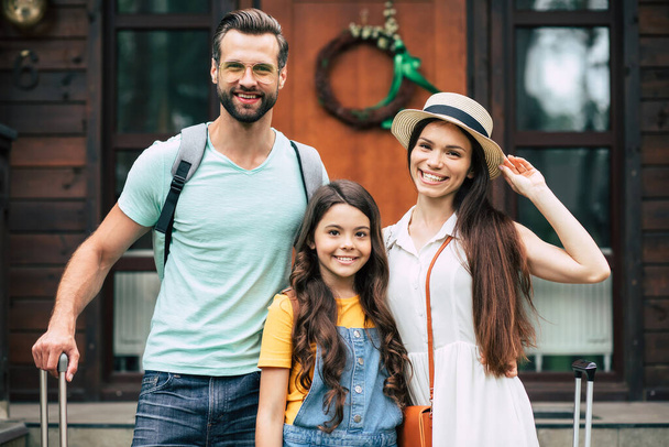 Photo of a beautiful young tourist family on vacation while them standing on forest hotel background and waiting to get settled in the room with backpacks and suitcases. - Foto, Imagen