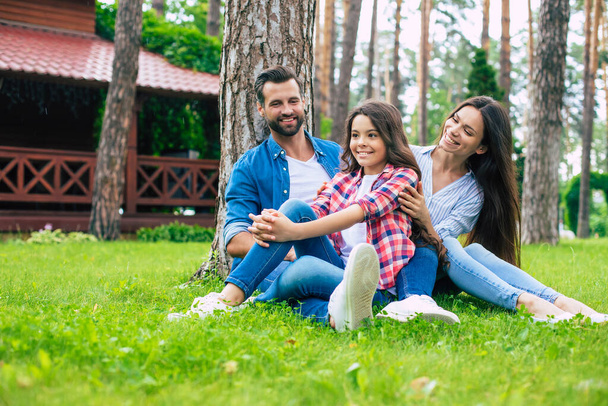 Beautiful happy family while sitting together on the grass and hugging each other, relaxing outdoors on suburban house background. - Φωτογραφία, εικόνα