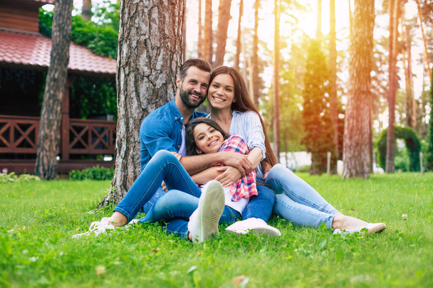 Beautiful happy family while sitting together on the grass and hugging each other, relaxing outdoors on suburban house background. - Photo, Image