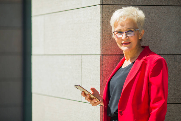 beautiful elderly woman in age with a short haircut and glasses in a red jacket with a phone - Foto, Imagem