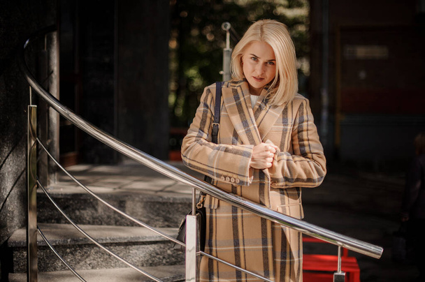 Young beautiful blond woman in plaid coat stands near metal handrail. - Valokuva, kuva