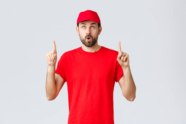 Online shopping, delivery during quarantine and takeaway concept. Impressed, curious employee in red uniform cap and t-shirt, open mouth interested, read banner, pointing fingers up - Photo, Image