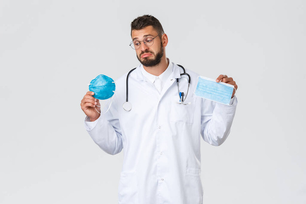 Gesundheitspersonal, Krankenversicherung, Pandemie und Covid-19-Konzept. Unsicherer Arzt in weißem Kittel, Brille und Stethoskop, schaut unentschlossen auf Beatmungsgerät, zeigt medizinische Masken und PSA - Foto, Bild