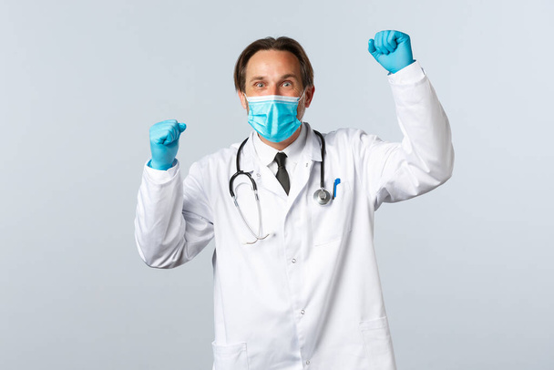 Covid-19, preventing virus, healthcare workers and vaccination concept. Relieved supportive doctor in medical mask and gloves, raising hands up as chanting, celebrating achievement of goal - Foto, Imagen