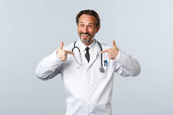 Ковід-19, спалах коронавірусу, медичні працівники та концепція пандемії. Щасливий професійний чоловік-лікар у білому пальто просуває власну клініку або послуги, вказуючи на себе, хвалиться
 - Фото, зображення