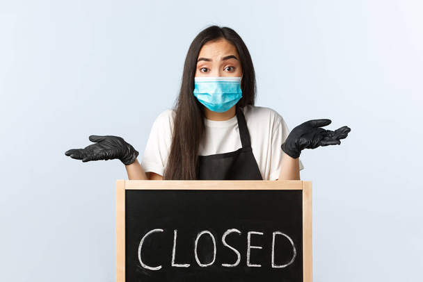 Small business, covid-19 pandemic, preventing virus and employees concept. Confused and puzzled asian female employee in medical mask shrugging, dont know when cafe opens after lockdown - Фото, изображение