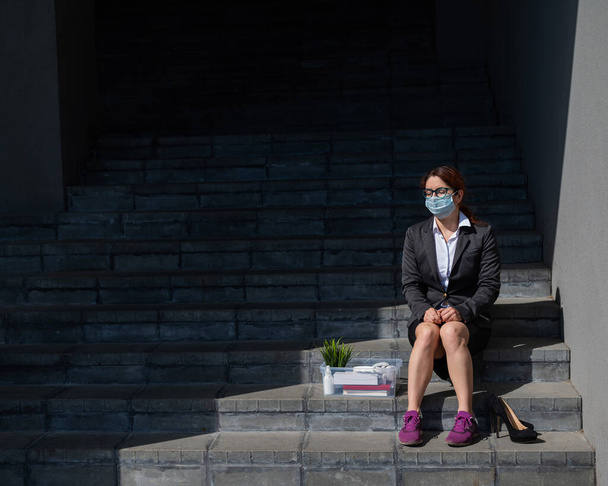 Unhappy woman in a mask sits with a box of personal stuff on the stairs outdoors. A female office employee was fired. The economic crisis at the height of the coronavirus epidemic. - Фото, зображення