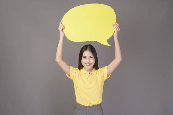 protrait of young confident beautiful woman wearing yellow shirt is holding a yellow empty speech on grey background studio - 写真・画像