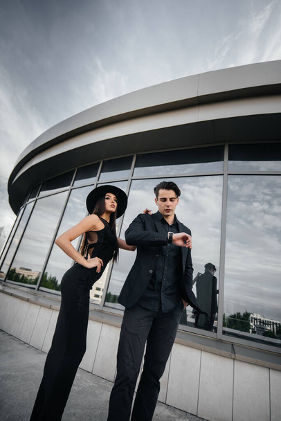 A beautiful, stylish pair of young people in black clothes and glasses stand against the background of an office building in the sunset. Fashion and style - Photo, Image
