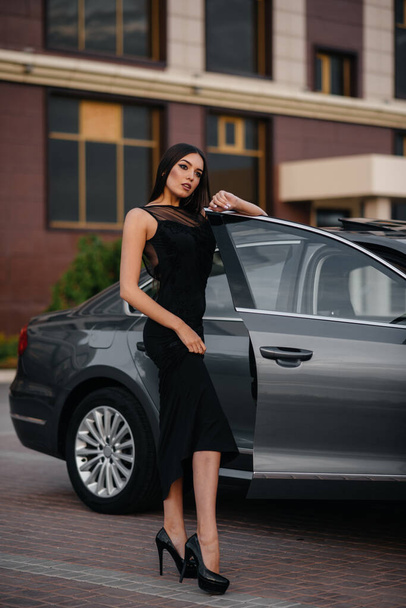 Stylish young girl stands near the car in a black dress. Business fashion and style - Fotografie, Obrázek